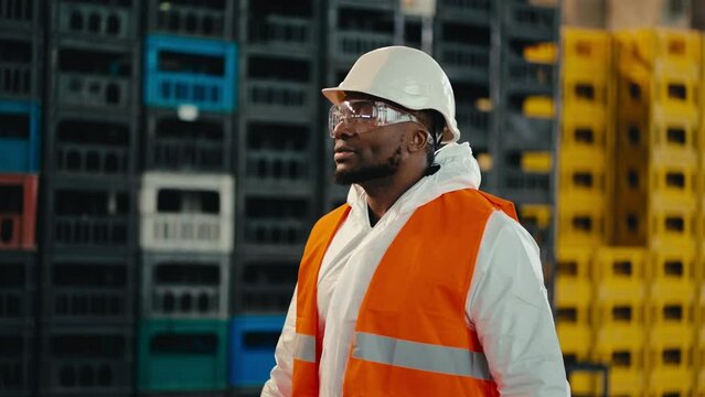A man with Black skin in a white protective uniform in an orange vest in transparent safety glasses walks along yellow and black boxes at a waste processing and sorting plant