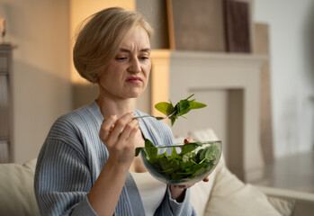 A middle-aged woman sits at home and with a dissatisfied look eats a green salad on a fork, which...