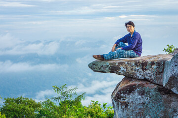 A handsome Asian man is sitting on a large rock that juts out from a steep cliff named Sut Phaen Din. Located at Pa Hin Ngam National Park in Thep Sathit district, Chaiyaphum province, Thailand.