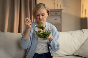 A woman is sitting on a beige sofa with a bowl of salad in her lap, looking dissatisfied as she eats. She is dressed in casual home attire with a light blue cardigan and is holding a fork mid-bite