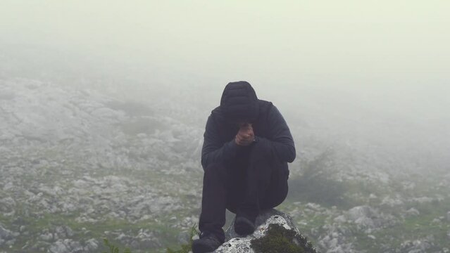 A man sits on a rocky mountain on a windy and foggy day as he tries to warm his hands, then gets up and moves on.
