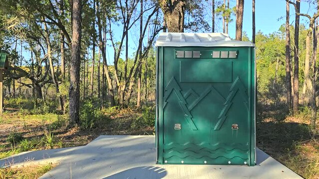 Pink inflatable axolotl entering and exiting portable toilet near woods viewed from the side