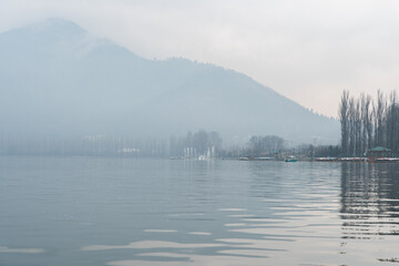 Misty Dal lake in Srinagar on the afternoon of January in Jammu and Kashmir. Still water with bare trees and shikaras in the background.