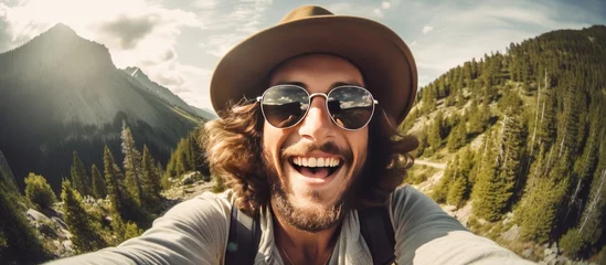 Gordijnen A smiling man wearing a hat and sunglasses is seen taking a selfie in the mountains. He appears cheerful and relaxed as he captures the moment. © TheWaterMeloonProjec