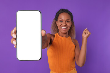 Young overjoyed beauty African American woman with phone in hands waves hand victoriously and looks at camera smiling advertising new website for online education stands in purple studio. White screen