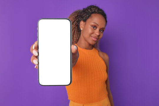 Young cute optimistic African American woman holding cellphone in hands and smiling modestly promoting mobile website or place to download music to smartphone stands on purple background. White screen