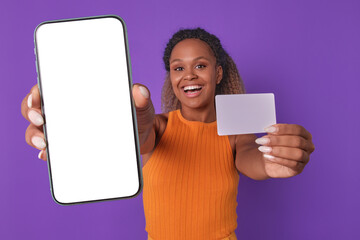 Young laughing beautiful African American woman with credit card and phone in hands urging to use...