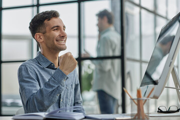 Happy businessman rejoicing success at workplace in office, looking at laptop screen with euphoric expression.