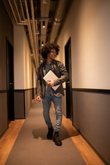 Curly-haired thin man with laptop walking in a hotel corridor