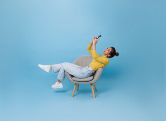 Happy young Asian teen woman using mobile phone. While her sitting relax on chair isolated on blue...