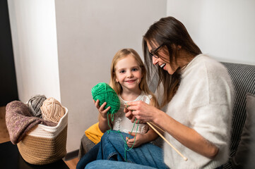 Mother teaching her daughter to knot on sofa in home interior knitting with her daughter