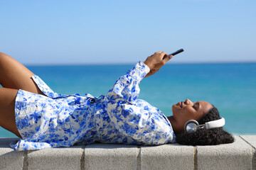 Happy black woman watching media lying on the beach - 755438084