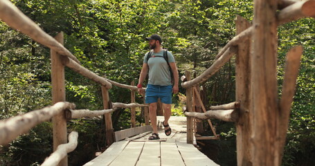 Fototapeta na wymiar A man in hiking gear with a backpack walks across an epic suspension bridge in a national park. A cinematic adventure of traveling in the mountains. A man walks through a small town on a mountain rive
