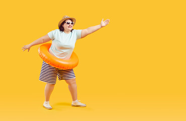 Fototapeta na wymiar Happy funny fat plus size woman in sunglasses with rubber ring pointing to the side with hand to copy space and having fun on studio yellow background. Vacation and travel concept.