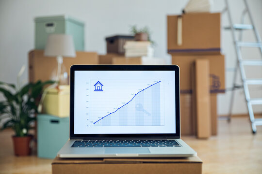 Rising line graph on laptop screen in front of cardboard boxes in an empty room in a new home