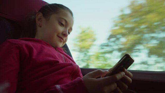 Portrait of cheerful smiling cute young little girl riding in public transport with mobile phone in bus. Cute child chatting with friends. Road trip automobile travel. Excursion. Child on train