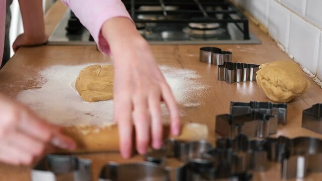 Children rolling out cookie dough with a wooden rolling pin on the kitchen countertop. Metal baking molds. Joy of childhood baking, family traditions, homemade treats. Siblings Helping to Mom together