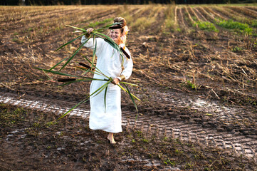 Happy woman in a dress dances barefoot on a muddy field, holding reeds in her hands
