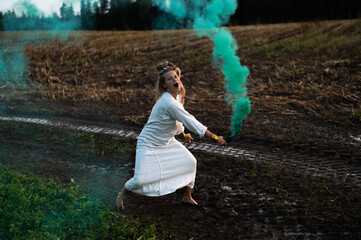 Happy woman in white dress with green smoke bomb dancing on muddy field
