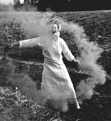 Happy woman in white dress with green smoke bomb dancing on muddy field