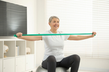 Fototapeta na wymiar Senior woman doing exercise with elastic resistance band on fitness ball at home