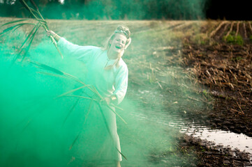 Happy woman in white dress with green smoke bomb and reeds dancing on muddy field