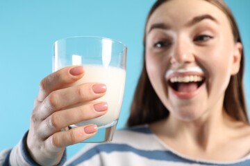 Emotional woman with milk mustache holding glass of tasty dairy drink on light blue background, selective focus