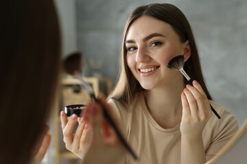 Smiling woman with freckles applying makeup near mirror indoors