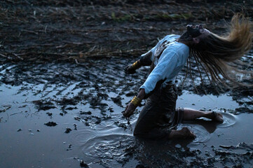 Dramatic portrait of a young woman kneeling in the black mud on field, cry of despair