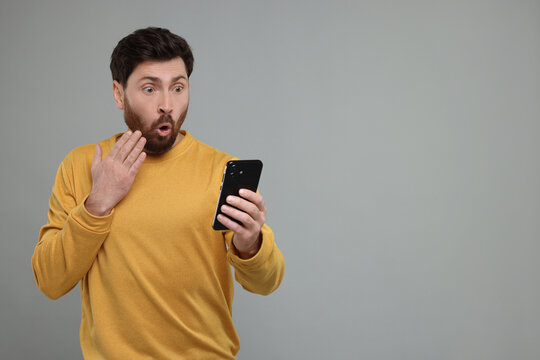 Surprised man with smartphone on grey background. Space for text