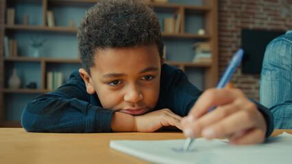 Sad upset lazy unmotivated tired exhausted fatigued little African American pupil schoolboy writing notes write homework task lying on table. Ethnic child boy kid son at desk boring class home lesson