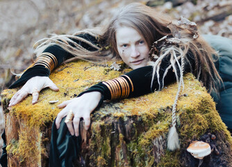 Female shaman in pristine forest nature laying on  tree stump, gold jewelry, shaggy hair