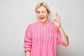 Smiling woman in pink sweater making OK sign with hand on a light background.