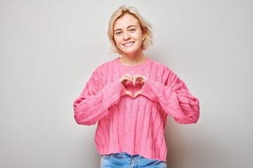 Smiling woman in pink sweater making heart shape with hands on light background.