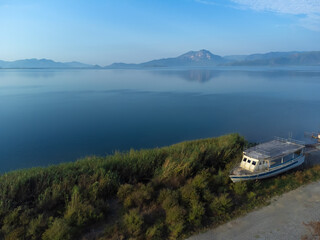 Drone view of old abandoned pleasure or fishing boat on shore of lake or sea overlooking mountains. Rusty and rotten boat washed ashore at dawn in sunshine. Fishing village in Turkey.