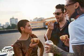 jolly attractive people with stylish sunglasses in vivid outfits enjoying hot dogs at rooftop party