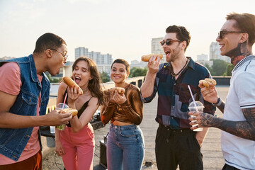 good looking jolly diverse friends in casual outfits eating tasty hot dogs at rooftop party