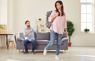 Young wife with packed backpack leaving her husband sitting on sofa at home. Unhappy couple man and...