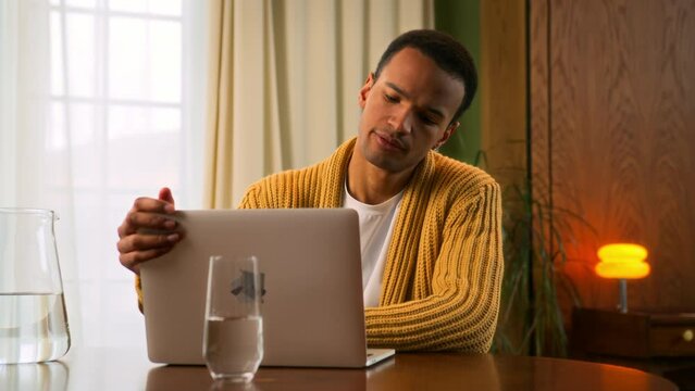 Young Latin man at laptop at home and smiling. gets disappointing news and becomes upset, closes computer to rethink and pause. male student of online education gets failure and feels frustrated
