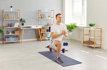Focused young man in activewear having a fitness workout at home, standing on a yoga mat in the living room, and doing a forward lunge exercise. Sport, wellness, physical activity concept - obrazy, fototapety, plakaty