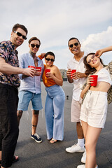 joyful multicultural friends in vibrant clothes holding red cups with drinks and smiling at camera