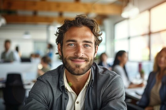 A handsome man with a genuine smile poses in a bustling open office space, wearing casual attire