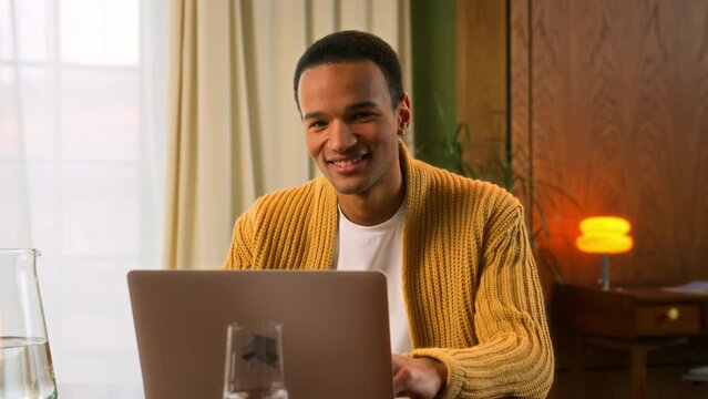 portrait of young Latin man sitting at table working at laptop at home, looks in camera nodding head and smiling in sign of assenting and success. contented and pleased student of online education