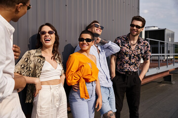 joyous multiracial friends in vivid clothing talking to each other happily and posing by wall