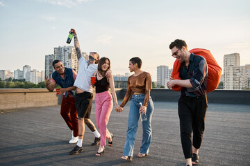 joyous multiracial friends with different drinks and bean bag chair in hands at rooftop party