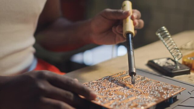 Cropped shot of unrecognizable Black male engineer in orange coverall pants and white undershirt soldering PCB at table in workshop