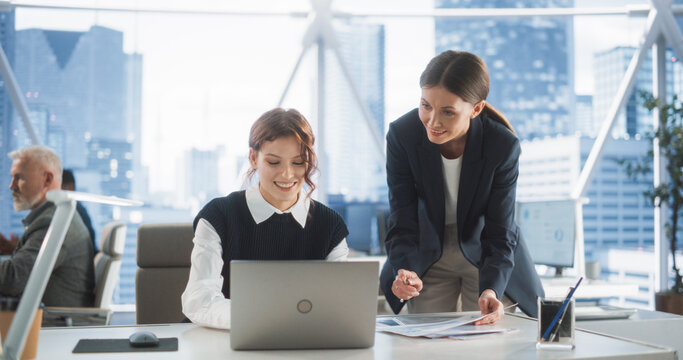 Professional Female Lawyer and Intern Talking And Working On Laptop Computer In Skyscraper Office In Downtown. Two Caucasian Women Studying New Case in Workspace Of Law Firm With Diverse Colleagues.