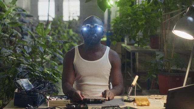 Waist up slowmo portrait of African American male engineer in white undershirt posing for camera with motherboard in hands wearing headband magnifying glasses with led lights