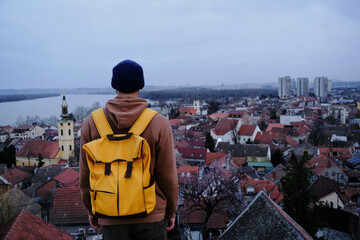 Danube river, tiled red roofs of houses and clock tower of church. A young guy backpacker travels and enjoys tourist spots. Belgrade, Serbia the old town Zemun. Rear view.