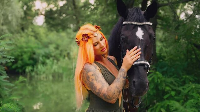 Real people happy fantasy woman with horse in green forest summer nature. Girl sexy fairy tale nymph hand holding reins of mare gently strokes muzzle fur pet animal. orange red long hair wreath. 4k